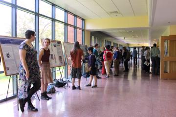 In the sunny Bent Corridor, people gather around research project posters on easels.