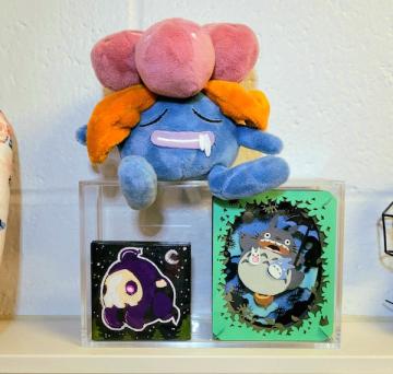 A display case filled with a paper theater of Studio Ghibli's "My Neighbor Totoro" and a 2x2 painting of a Pokémon, Duskull. On top of the display case sits a stuffed plush of another Pokémon, Gloom.