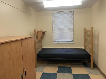 The picture shows one half of a bare divided double dorm room. At the back of the room, a bare blue mattress lays on top of its bed frame and a cabinet is placed on the left wall. No decorations or belongings are inside of the room.