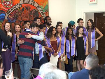 A group of students wearing purple stoles stands in front of an illustration with the words solidarity, peace, progress.