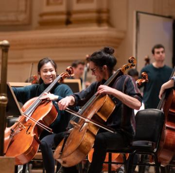 Two students playing the cello in an orchestra.