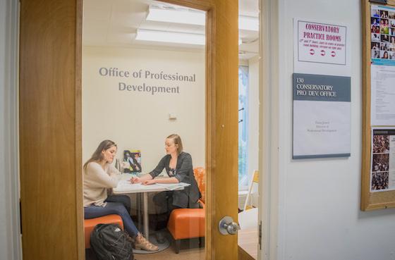 Two people have a discussion at a small table behind a glass door marked 'Office of Professional Development'