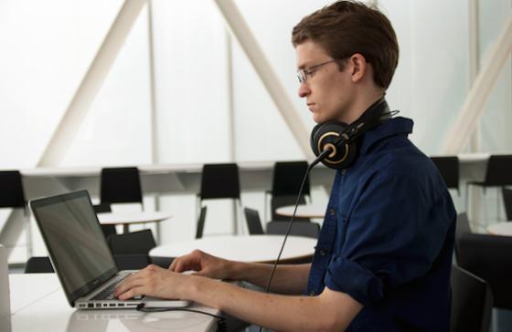 A student works on a laptop computer at a table, a pair of headphones resting around his neck
