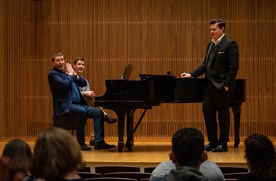 two men sitting at the piano with a student vocalist standing on stage