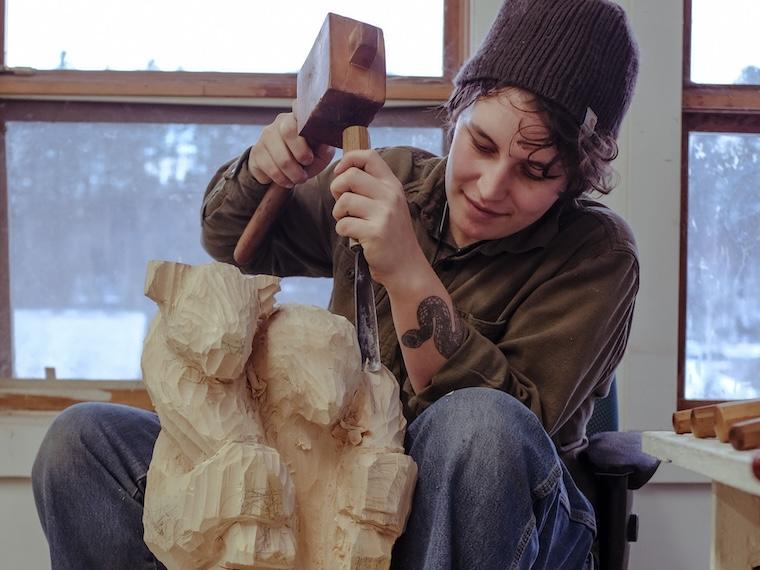 Eva Sturm-Gross carving a wooden sculpture with a mallet and chisel.