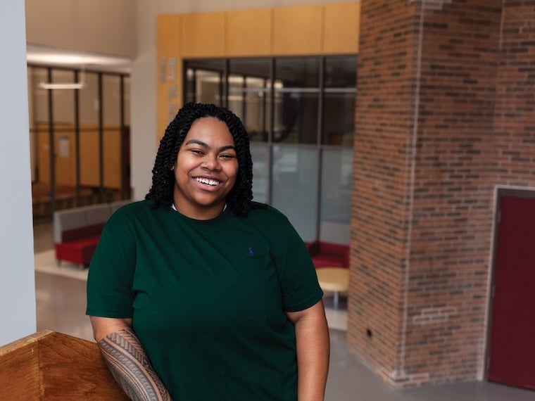 A smiling student with long dark hair wearing a dark green shirt stands indoors near a wooden railing. The background shows a brick wall, glass panels, and red seating in a modern building.