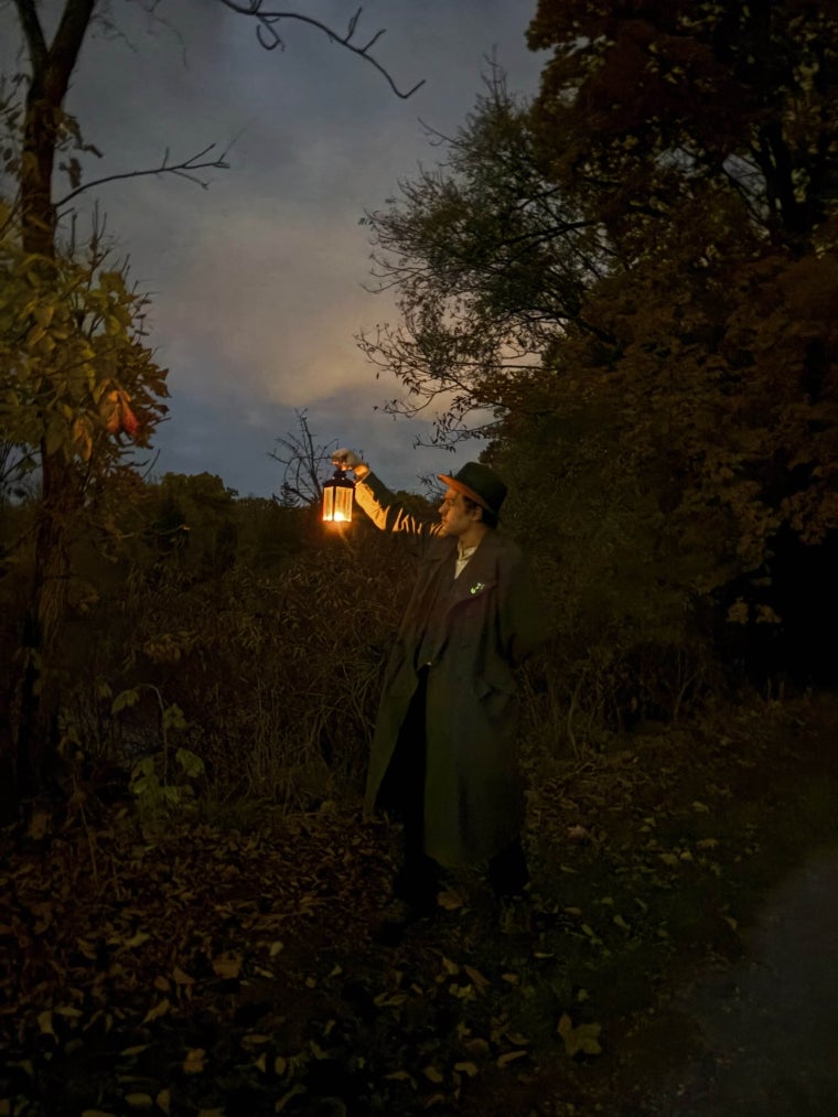 A figure in a coat stands in the Oberlin Arboretum at night, holding a lantern.
