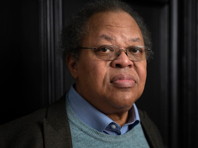 man with dark curly hair and glasses, wearing a blue sweater and dark jacket
