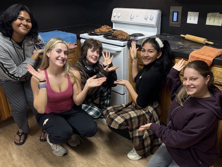 Five students kneeling around a small dishwasher in a kitchen, smiling and waving at the camera.
