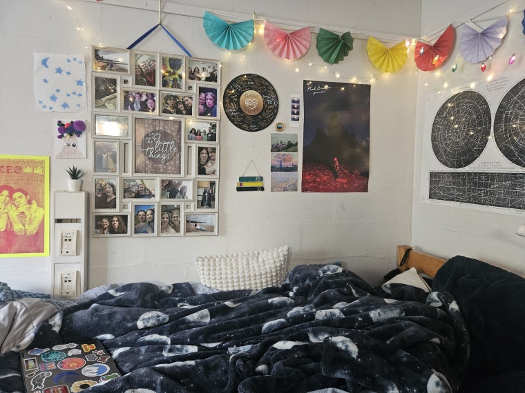 Part of a dorm room, with posters, photos, and colorful banners on the walls and throw pillows and a moon-patterned blanket on the bed.