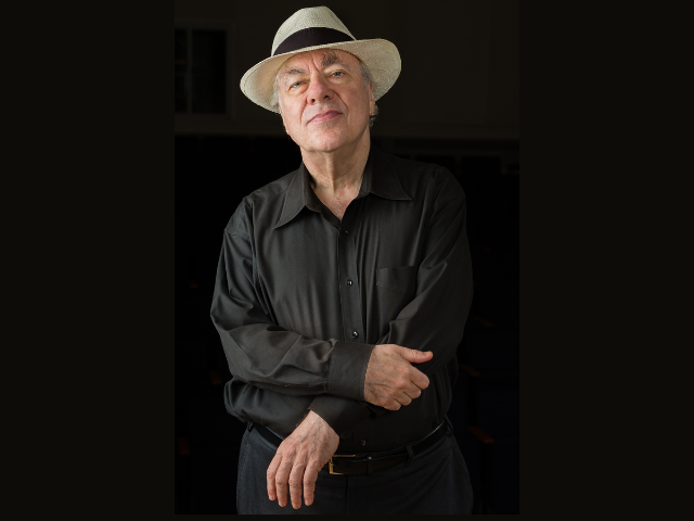 a man wearing black clothes and a white hat with a black band, standing and slightly smiling at the camera