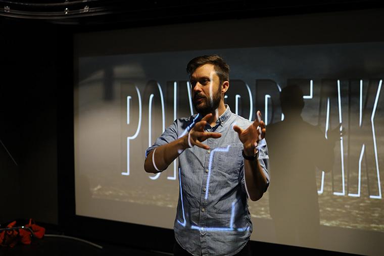 A professor speaks to a class while standing in front of a movie screen. An image is projected on his face and shirt.