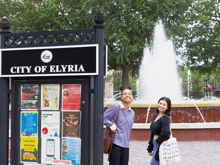 Two students in front of a public fountain. A sign says City of Elyria.