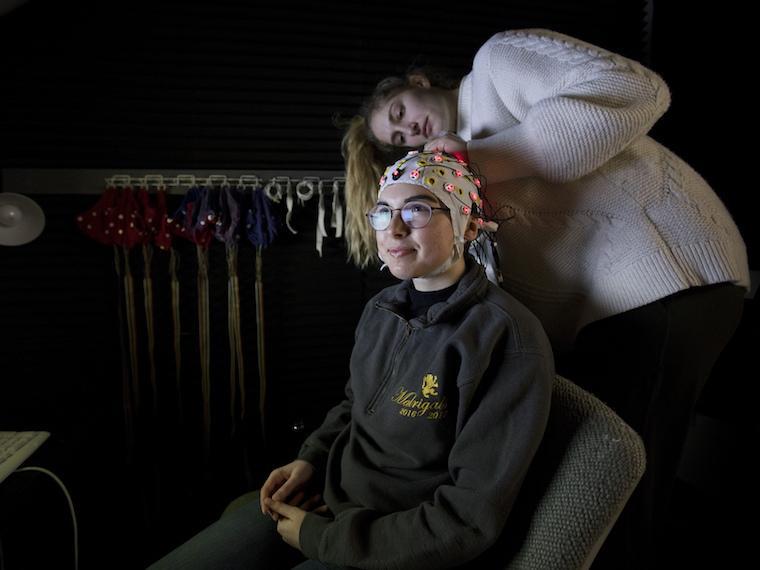 A in a white sweater adjusts an EEG cap on a student, who is seated in an office chair in front of a computer keyboard. The cap is off-white with pink and yellow nodes and wires coming out of it.