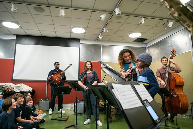 An Oberlin string quartet performs in front of a group of children. One of the musicians holds up a microphone to one of the children. They are smiling.