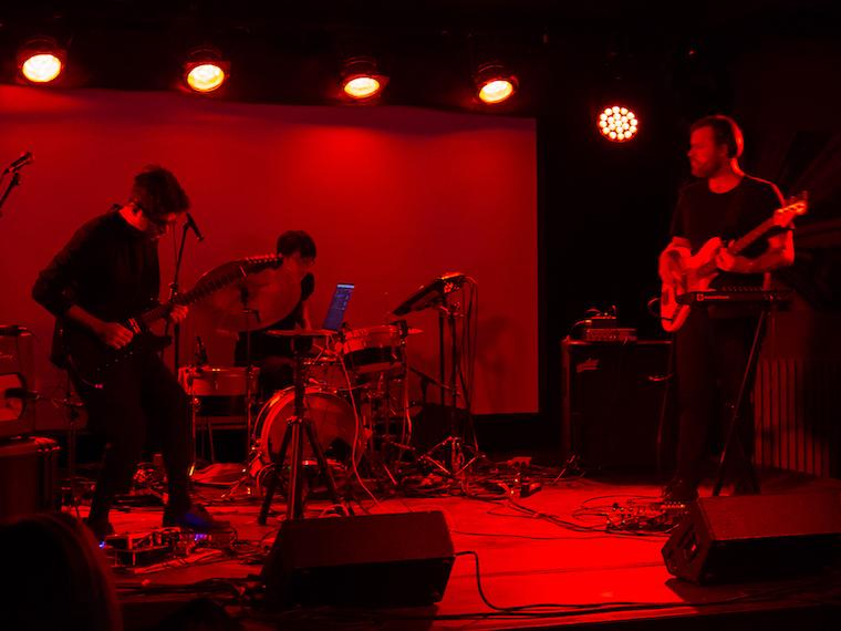 A rock trio (guitar, bass, and drums) performs under red stage lights.