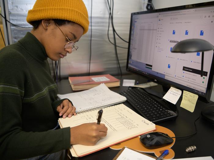 A student carefully writes data in a log book while reviewing data at a computer.