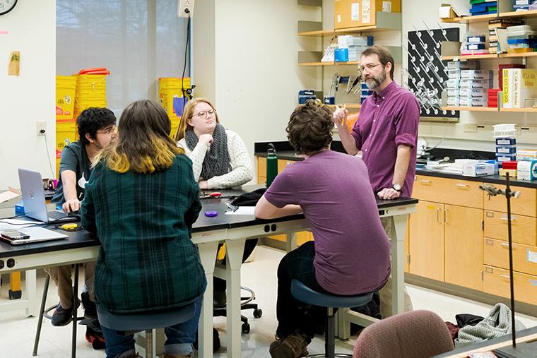 Four students and a professor have a conversation at a lab table.