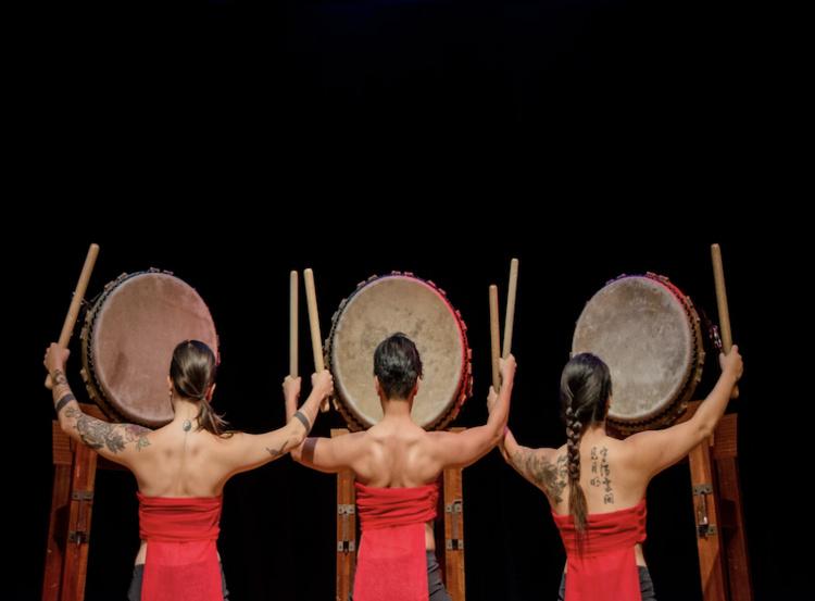 Three drummers face chu with arms outstretched.