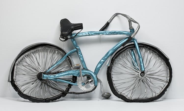 A soft sculpture depicts a realistic light blue bicycle that droops and sags due to the material’s nature, with long threads along the seams of the fabric.