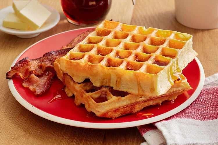 Two strips of bacon next to two stacked square waffles on a red plate.