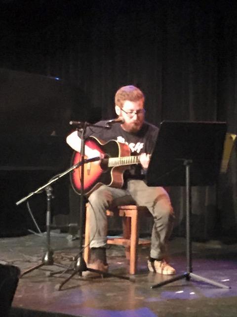 A guitarist is seated by microphones and a music stand