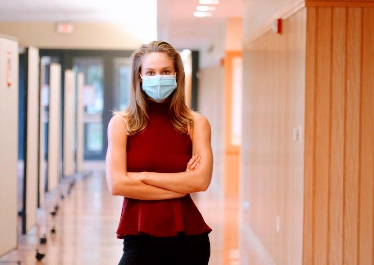 A girl wearing a surgical mask stands with arms folded in a hallway.