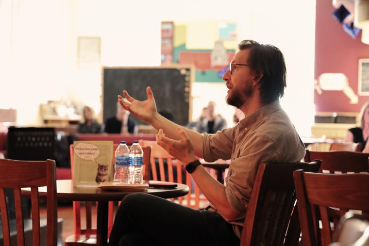 Chris Eldridge, seated at a cafe table, gesturing towards a music ensemble off-frame.