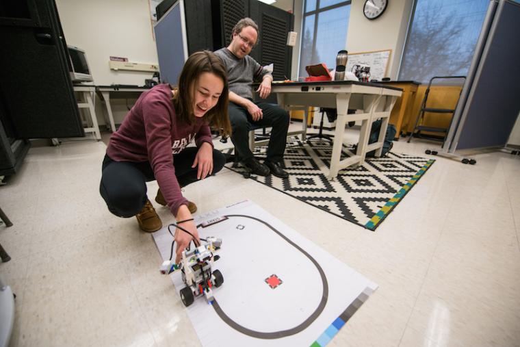 A student positions a small device with wheels on a track while a professor looks on