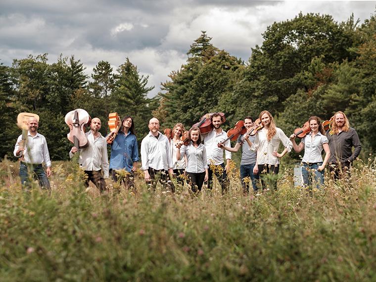 The ACRONYM Ensemble standing in a sunlit field holding Baroque instruments.