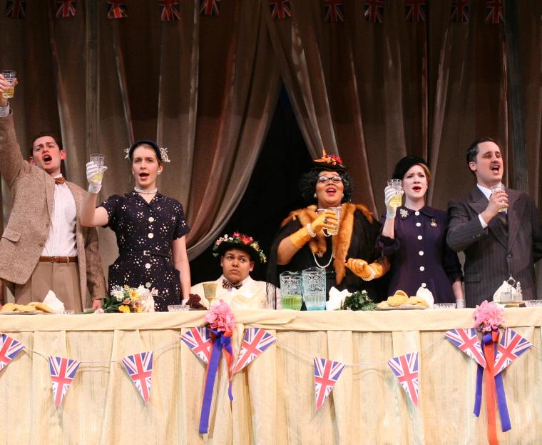 In a scene from the production, performers stand at a long table with glasses raised.