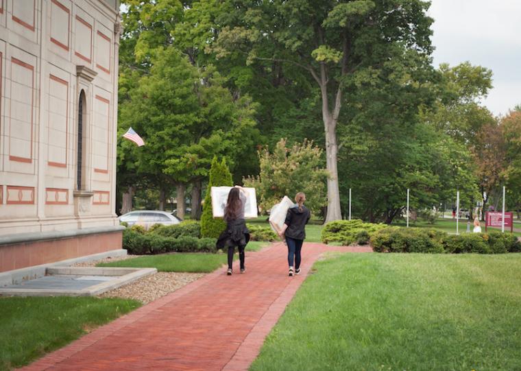Two students walk away from the museum, each carrying a framed artwork.