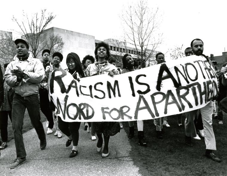 Protesters in front of the main library carry a banner that says Racism is Another Word for Apartheid.