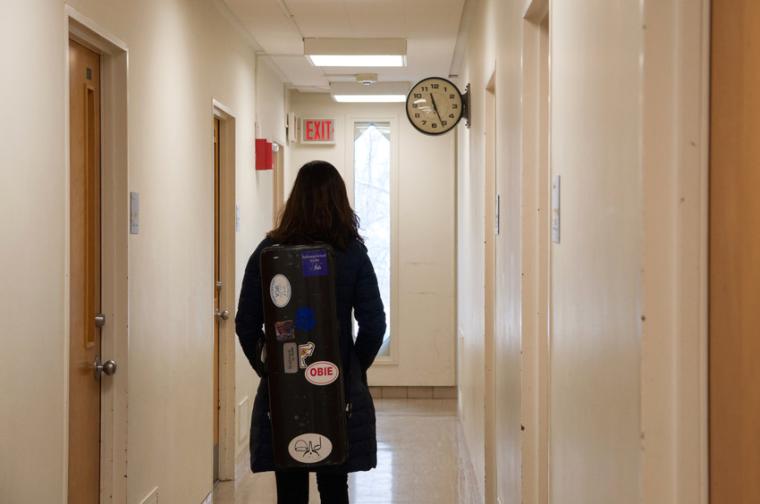 A student walks down a hallway, carrying an instrument case covered in stickers.