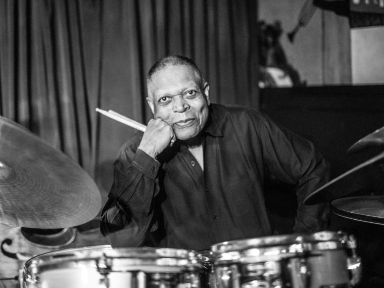 man sitting at drum kit with drumsticks in hand smiling at camera