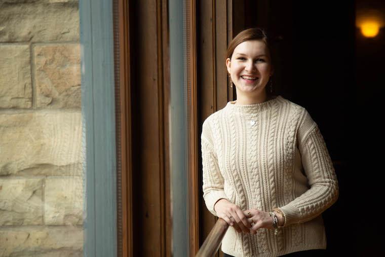 Picture of woman in beige sweater standing by a window.