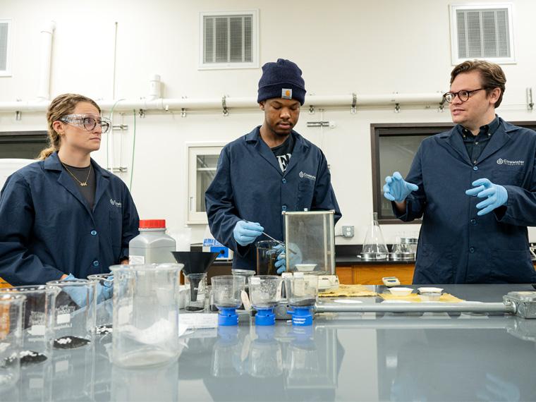 Two students wearing protective gear work in a lab with an employee