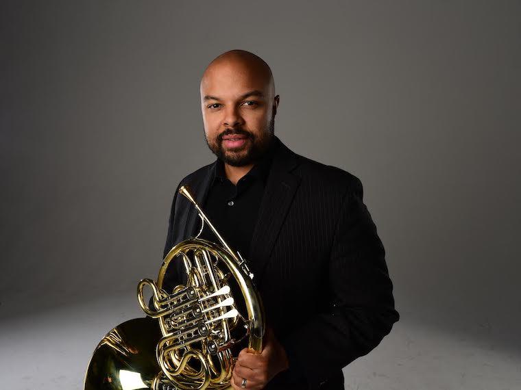 Man seated, wearing concert black, holding a french horn