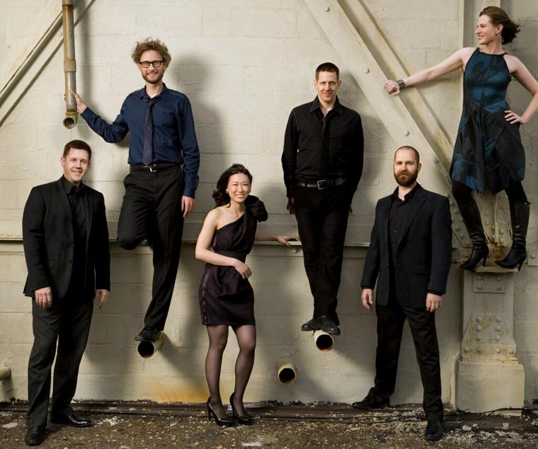 The six musicians pose in concert attire against a painted, industrial brick wall. Three of them are standing on pipes or a beam emerging from the wall about 2 or 3 feet above the floor.
