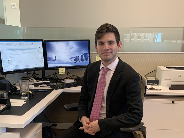 man wearing suit and tie sits at a desk in office space.