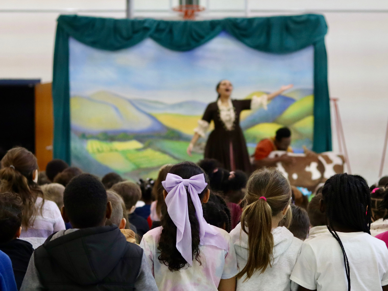 photo of young children sitting in a school gymnasium, facing a stage with a woman in a gown who is singing