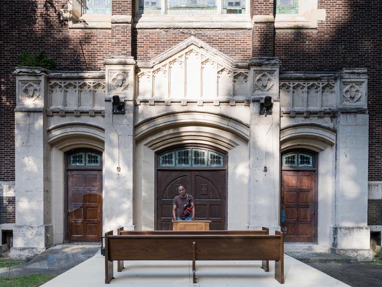 Outside the front doors of a church, the artist stands behind a pulpit and faces two wooden pews.