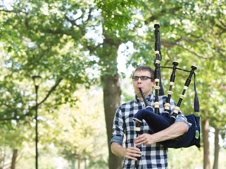 Man in plaid shirt playing bagpipe in a leafy setting