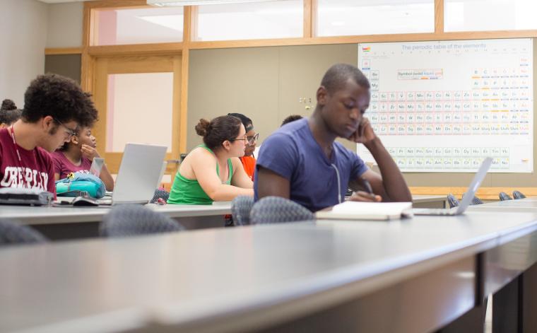 students sitting at long tables with computers and laptops in a classroom