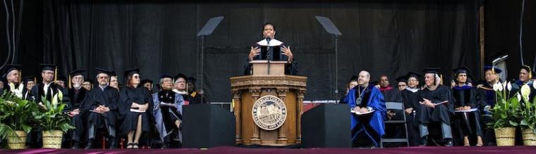 Michelle Obama speaks at a podium bearing the Oberlin College seal. Dignitaries in academic robes are seated on both sides.