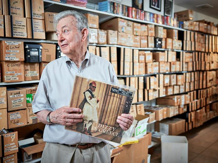 Neumann holds an LP, Dizzy in Greece. Behind him is an endless shelf filled to the ceiling with labelled boxes.