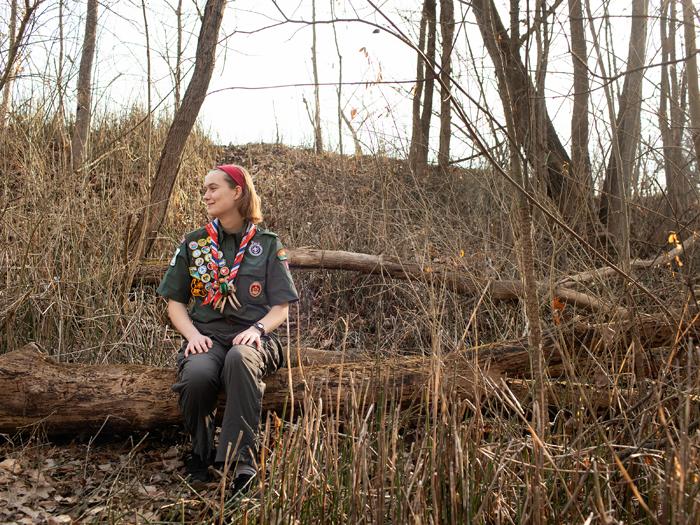 Eagle Scout Nissa Berle sitting on log in wooded field.