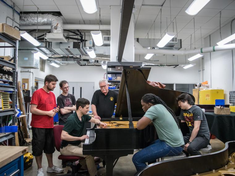 Teacher and students gathered around a Steinway piano in a repair shop.
