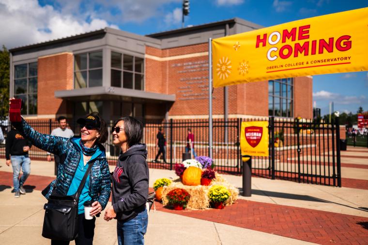 Two women standing before a football field and a Homecoming banner taking a selfie