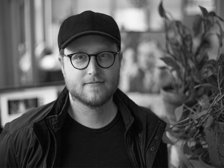 a person with glasses wears a baseball cap and a dark shirt and jacket